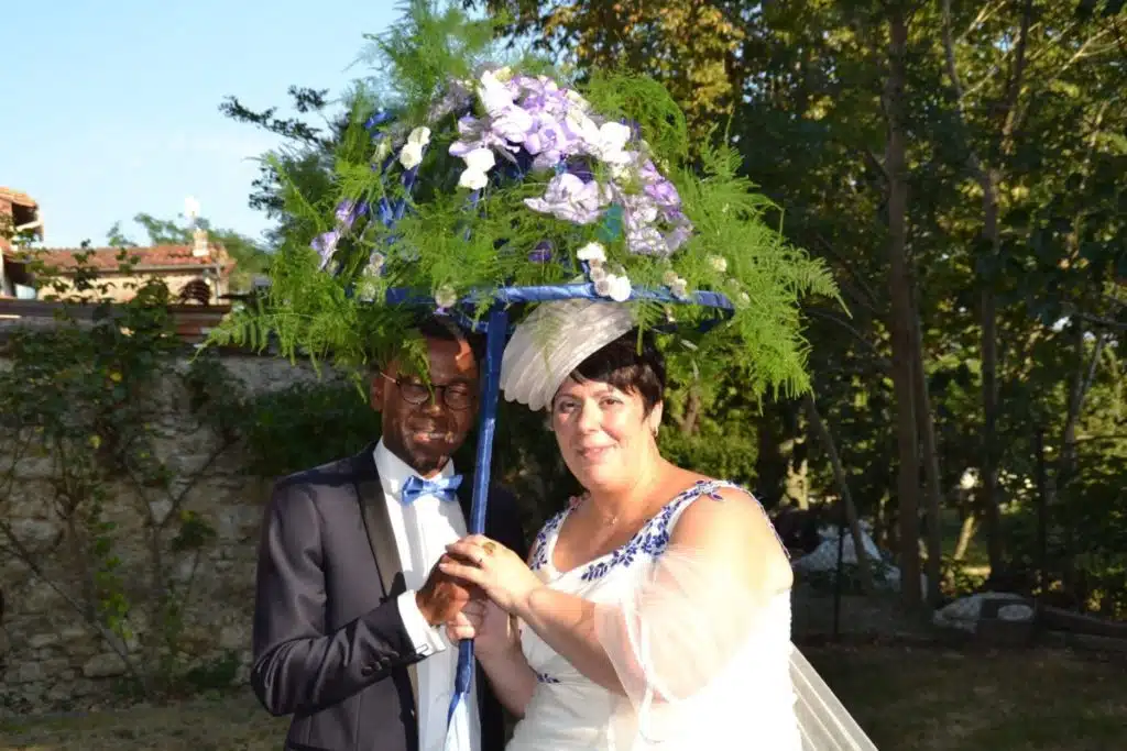 Couple de mariés tenant un parapluie décoré de feuillages et d’orchidées violettes lors d’une séance photo en extérieur.
