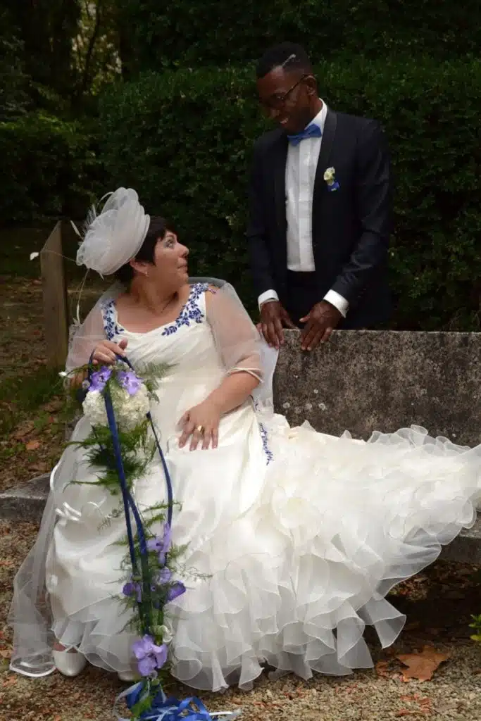 Mariée assise tenant un bouquet retombant composé d’orchidées violettes, de feuillages et de fleurs blanches, lors d’une séance photo en extérieur.