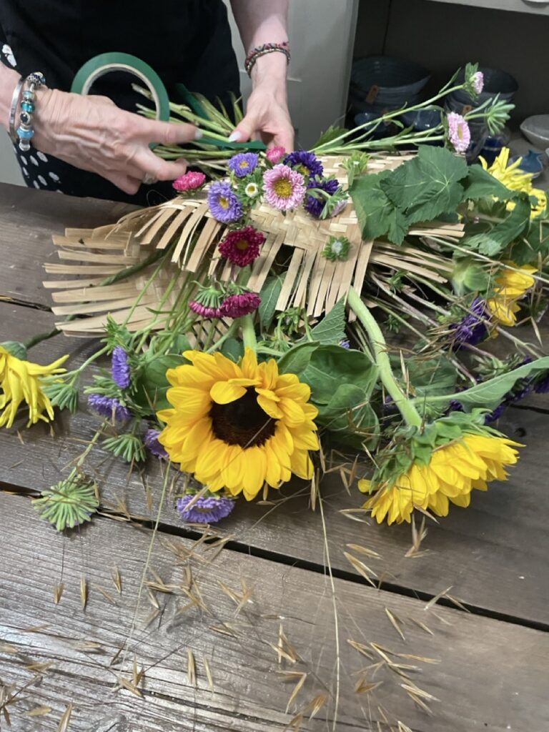 Participant composant une création florale avec des tournesols, des fleurs de saison et des matières naturelles lors d'un atelier convivial animé par Yann-H2o, artisan fleuriste créateur à Paris 11. Chaque atelier invite à explorer sa créativité dans une ambiance chaleureuse et bienveillante.