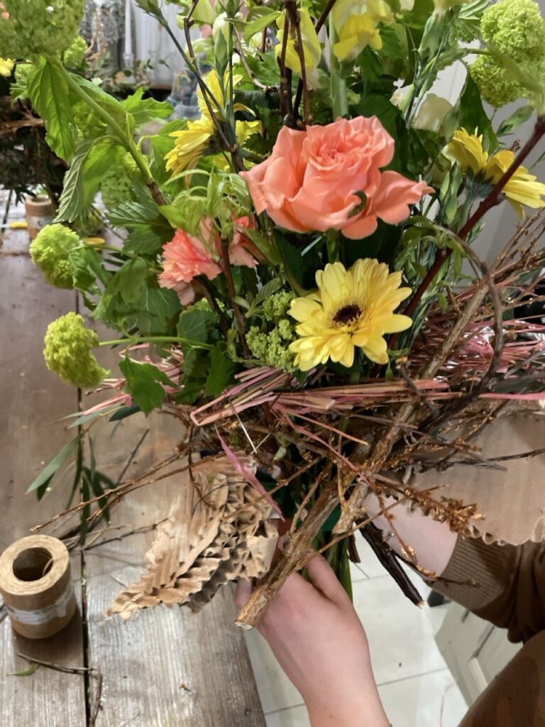 Bouquet structuré d’automne aux roses saumonées, gerberas jaunes et fleurs vertes, entouré de branchages naturels, créé lors d’un atelier floral à Paris 11 chez Yann-H2o.