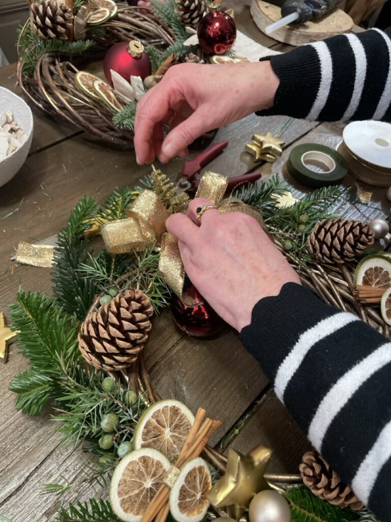 Une participante ajoute un ruban doré à sa couronne de Noël ornée de sapin, pommes de pin, oranges séchées et boules rouges, lors d’un atelier floral à Paris 11 chez Yann-H2o.