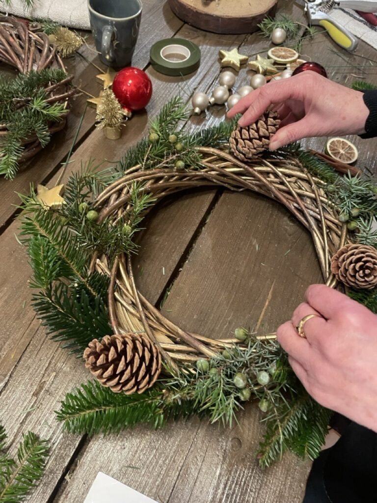 Une participante dispose une pomme de pin sur une couronne de Noël en branches tressées et feuillages de sapin, lors d’un atelier floral à Paris 11 chez Yann-H2o.