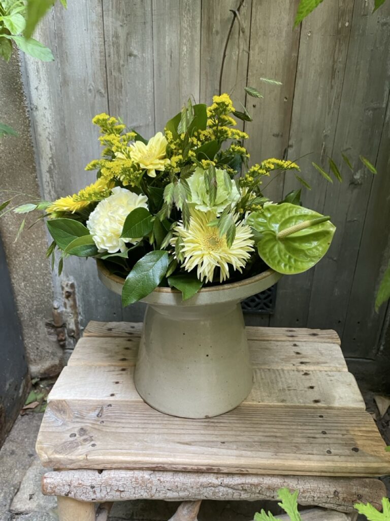 Composition de fleurs jaunes, blanches et vertes avec gerberas, œillets et anthurium, présentée dans un vase beige sur une table en bois.