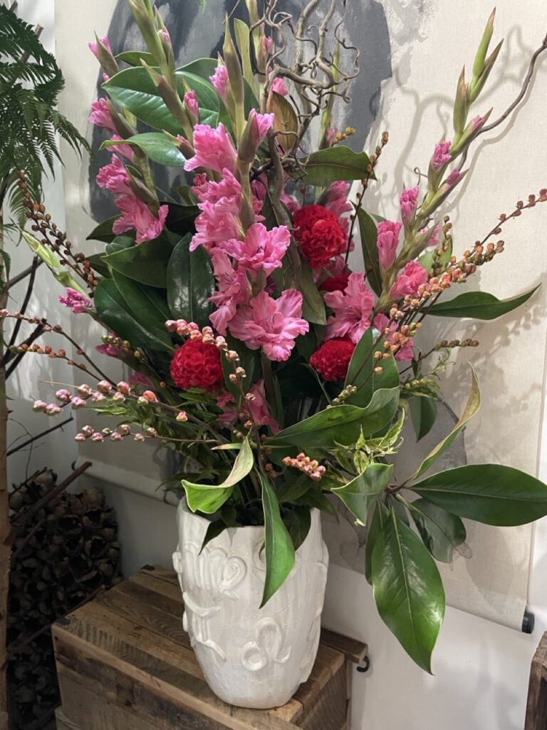 Composition florale de célosies fuchsia et glaïeuls roses, accompagnés de branches et de feuillages, dans un vase blanc sculpté chez Yann-H2o à Paris 11.